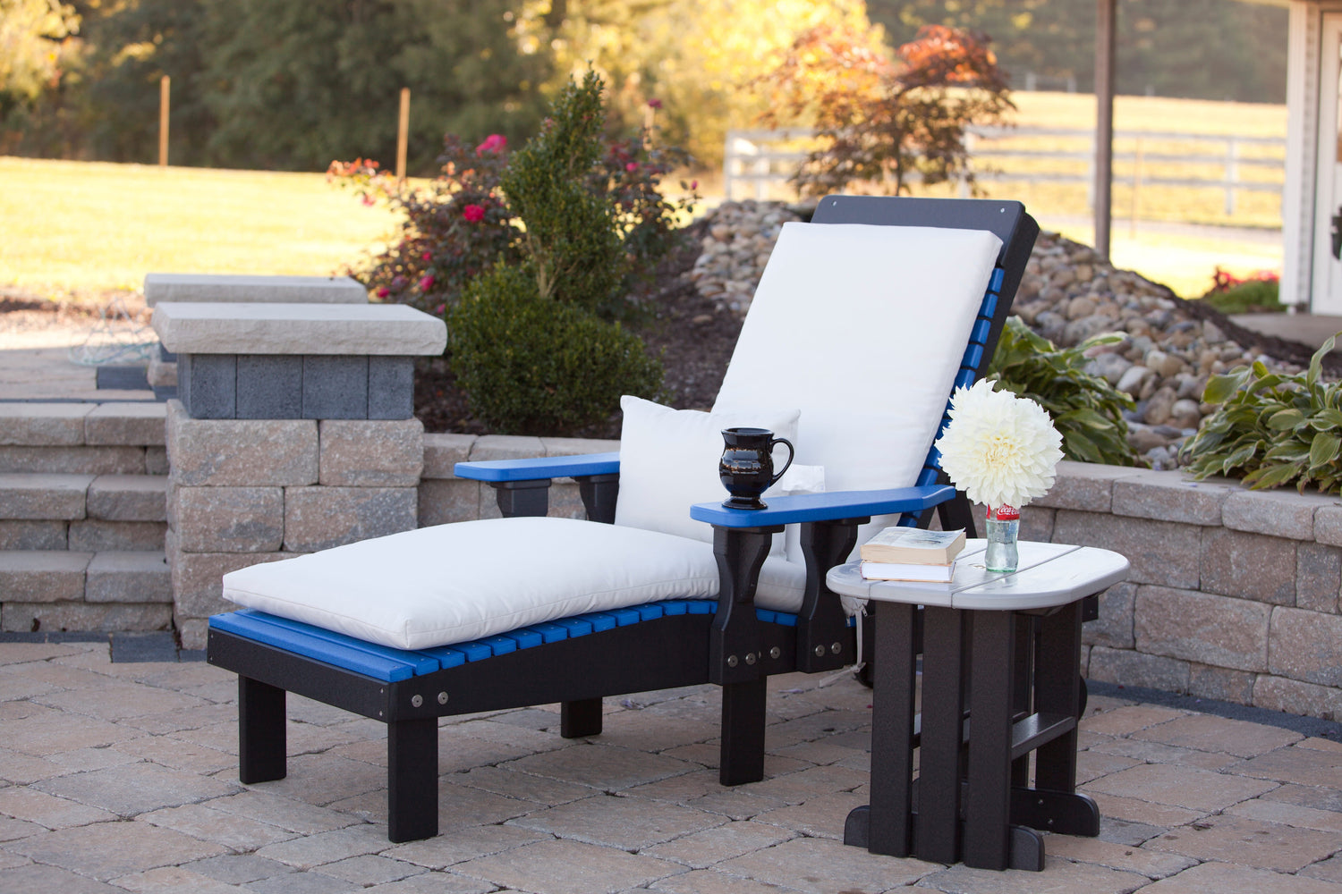 A blue and black outdoor chaise lounge chair with white cushions on a stone deck next to an end table.