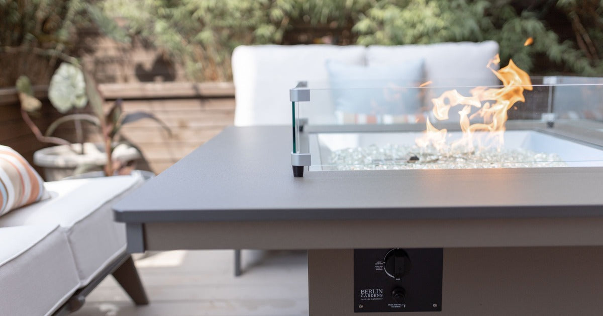 A close-up of the edge of an outdoor fire pit with glass beads, with cushioned couches and chairs in the background.