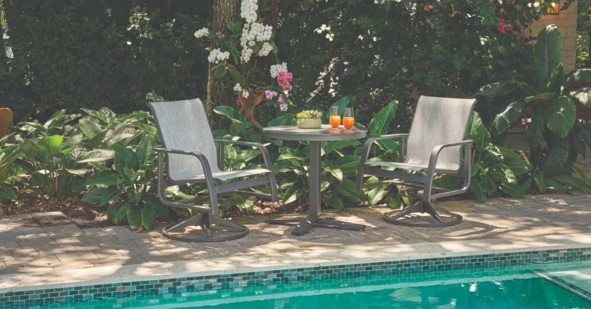 Two swivel lounge chairs on a poolside patio with a small table between them, with two glasses on it.