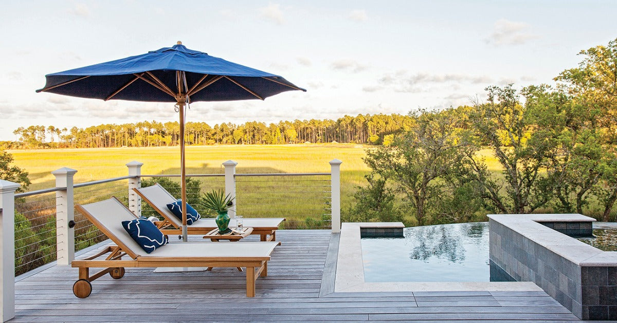 The backyard deck of a home with two chaise lounge seats with an umbrella between them next to a pool.