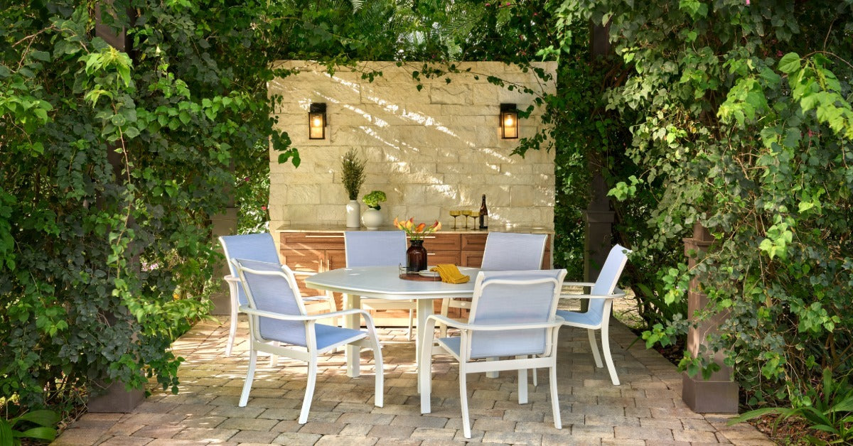 A stone patio in a backyard with vines and bushes growing on a pergola over a circular table with five chairs.