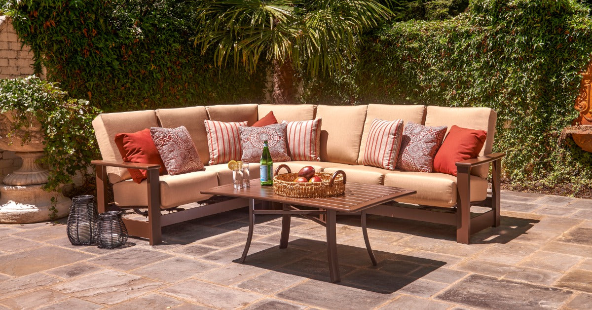 An outdoor sectional sofa on a stone-tile surface with beige cushions, red and grey throw pillows, and a coffee table.