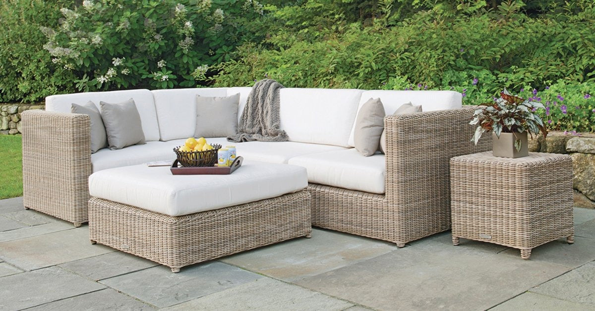 A wicker outdoor sectional with cream cushions on a stone patio beside plants, flowers, and green shrubs.