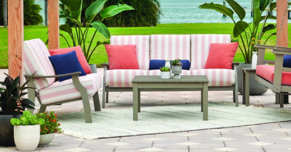 An outdoor patio seating area with a waterfront lawn view, with striped pink-and-white cushioned chairs, a loveseat accented by coral and navy throw pillows, and a coffee table on a rug.